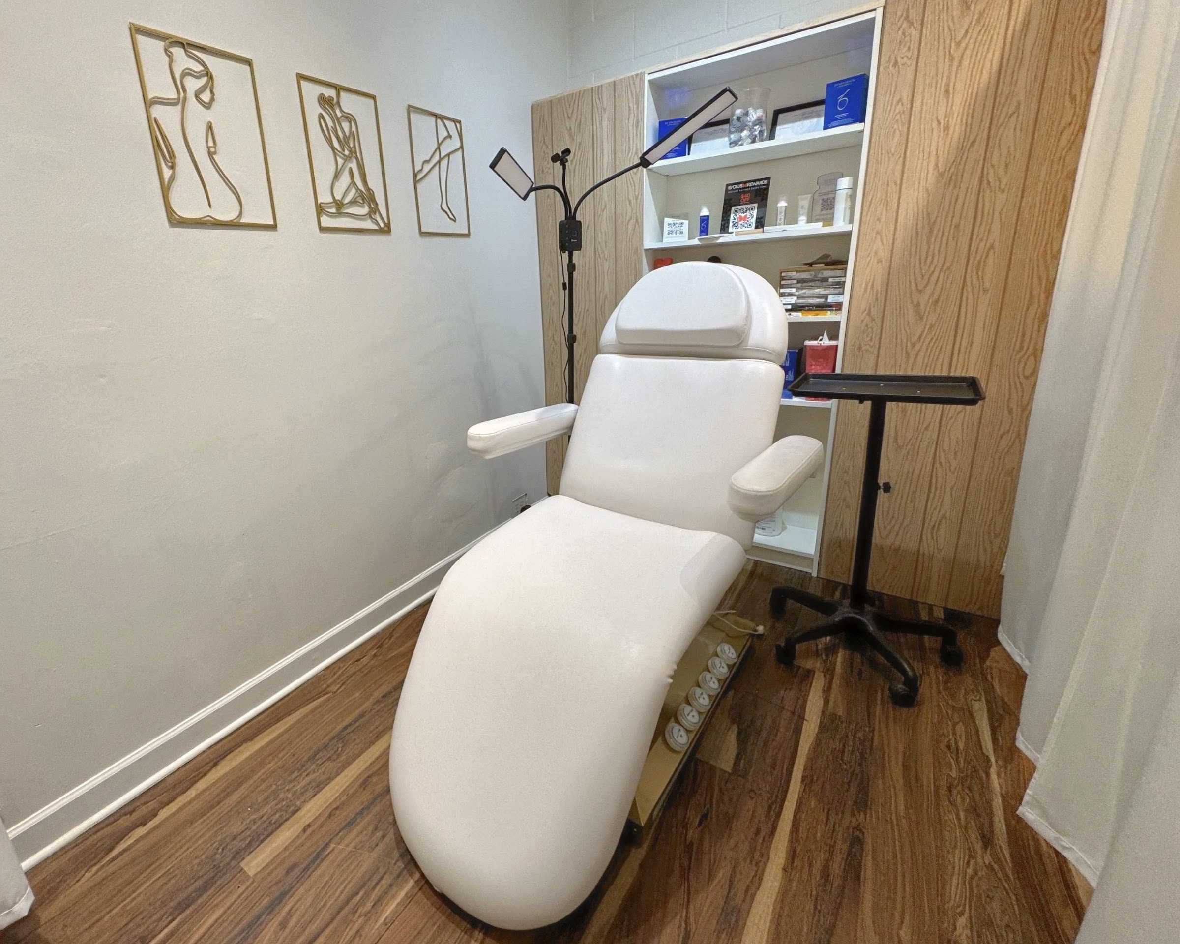 The treatment room at Defined Skin Spa — a white facial chair beside gold-framed line-art prints, warm wood floors and soft natural light